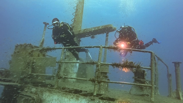 Two RAF Brize Norton Sub Aqua Club scuba divers navigating near the algae-covered metal railings of a sunken shipwreck in murky blue ocean water.